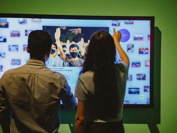 Freiheit 5_Isabel Noack.jpg Zwei Personen interagieren mit einem großen, wandmontierten Touchscreen in einem Ausstellungsraum.