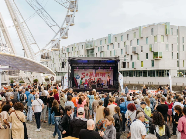 Hafensound beim Open Air Sommer Köln Ein lebhaftes Open-Air-Konzert in Kölns Hafengegend mit einer großen Menge, die eine Sommerbühne umringt.