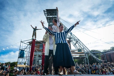 Dyptik Mirage beim Open Air Sommer Köln Eine bunte Gruppe von Akrobaten führt vor einem Kran in einer lebhaften Festivalumgebung eine spektakuläre Show vor zahlreichen Zuschauenden auf.