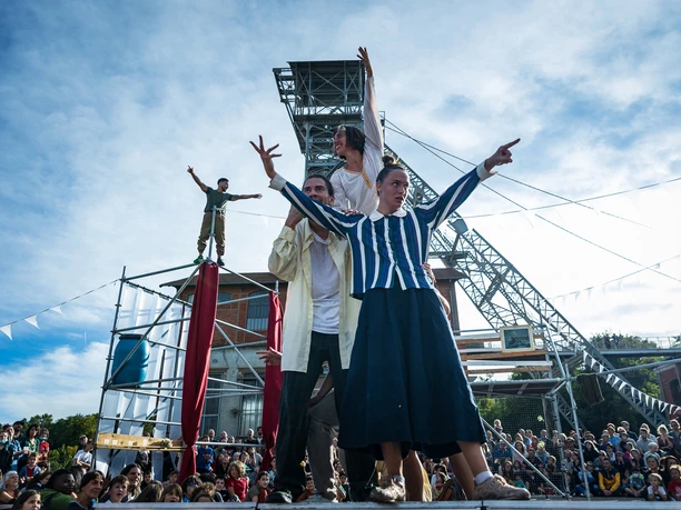 Dyptik Mirage beim Open Air Sommer Köln Eine bunte Gruppe von Akrobaten führt vor einem Kran in einer lebhaften Festivalumgebung eine spektakuläre Show vor zahlreichen Zuschauenden auf.