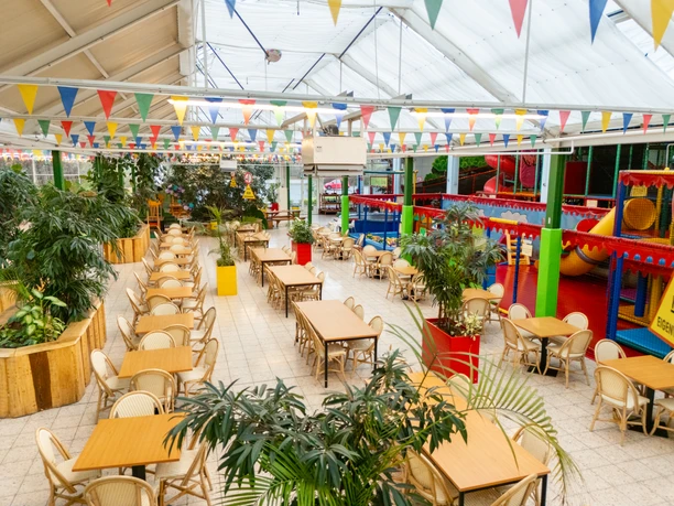 Große Indoor-Spielhalle mit bunten Spielgeräten, langen Tischreihen und farbenfrohen Wimpeln.
