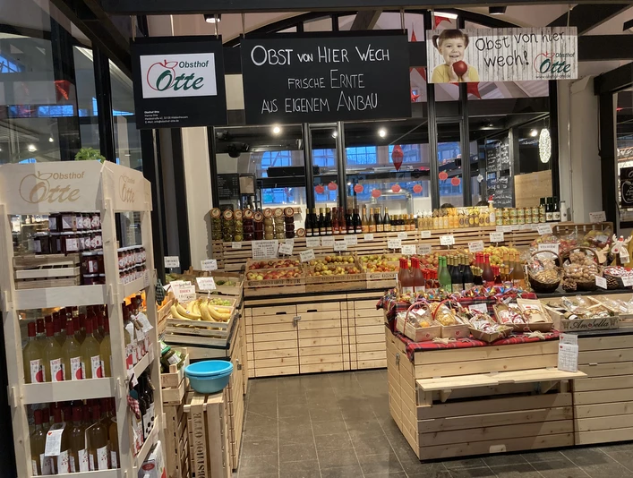 Häppchentour Markthalle_Otte (Maya Regtmeier 21).jpeg Ein Verkaufsstand in einer Markthalle präsentiert frisches Obst, Gemüse und regionale Produkte.