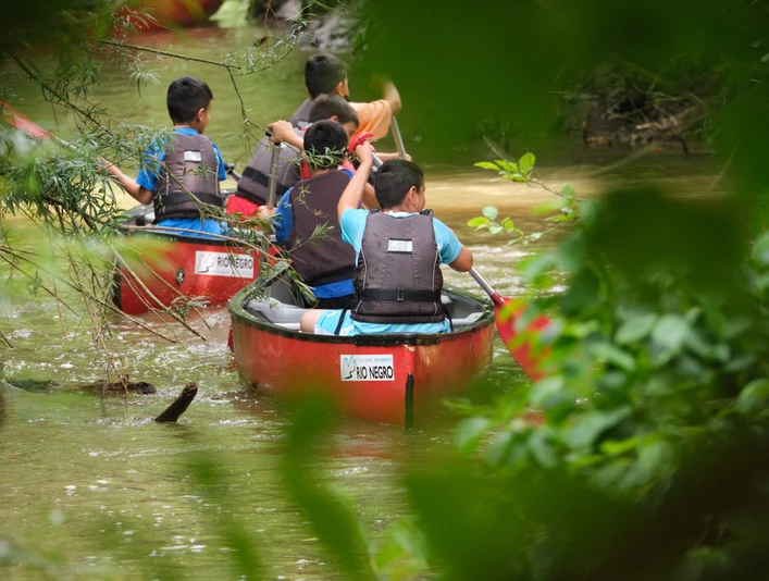 Kanufahren mit Rio Negro - Olaf Brandmeyer 2.jpg Vier Kinder in roten Kanus paddeln durch einen dichten, grünen Flussabschnitt im Wald.