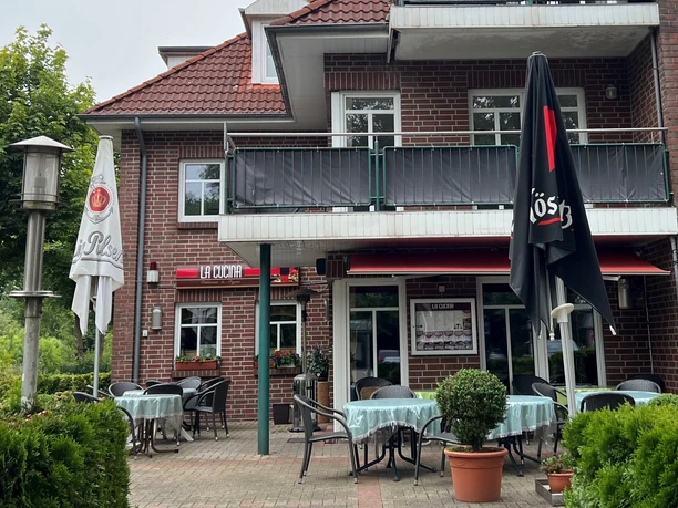 LaCucina2.jpg Das Bild zeigt ein Restaurantgebäude aus rotem Backstein mit weißem Fensterrahmen und einem roten Dach. Vor dem Gebäude befinden sich mehrere runde Tische mit blauen Tischdecken, umgeben von grünen Pflanzen und Sonnenschirmen. Die Terrasse ist einladend gestaltet und bietet eine freundliche Atmosphäre für Gäste.