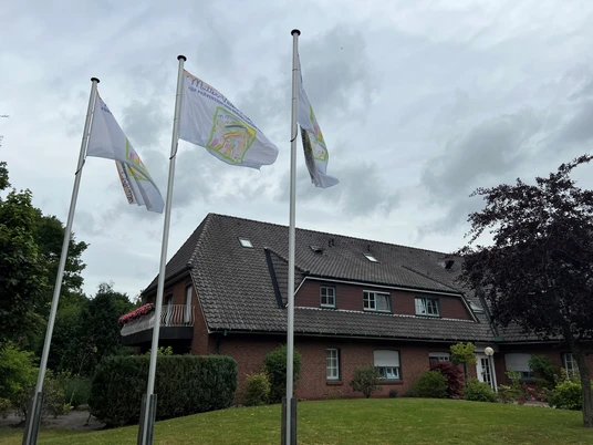 Außenansicht.jpg Gemütliches Backsteinhäuser mit drei wehenden Flaggen, umgeben von gepflegtem Grün unter grauem Himmel.