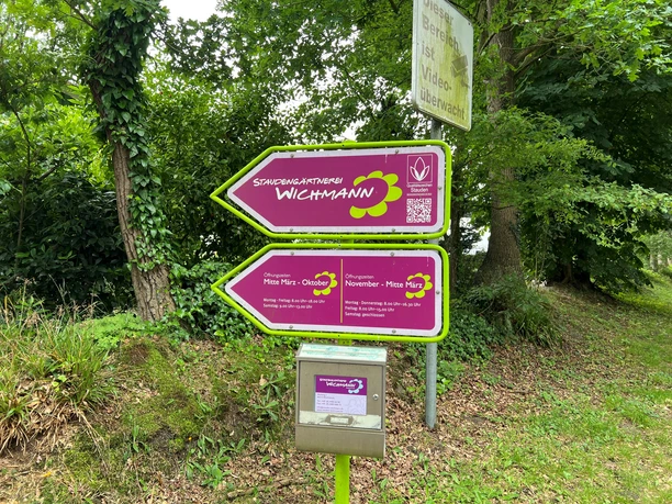 Schild.jpg Es ist ein Schild zu sehen, welches mit dem Logo von "Staudengärtnerei Wichelmann GmbH & Co.KG" in der Farbe lila ausgewiesen ist.