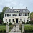 Bild5.jpg Ein großes, weißes Herrenhaus mit grauem Walmdach steht in einem gepflegten Garten hinter einem schmiedeeisernen Tor.