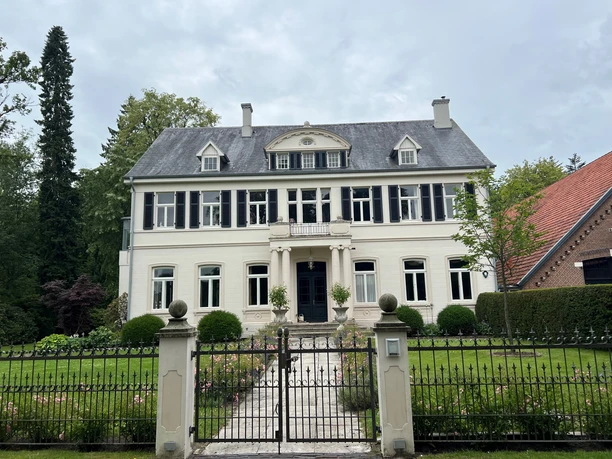 Bild5.jpg Ein großes, weißes Herrenhaus mit grauem Walmdach steht in einem gepflegten Garten hinter einem schmiedeeisernen Tor.
