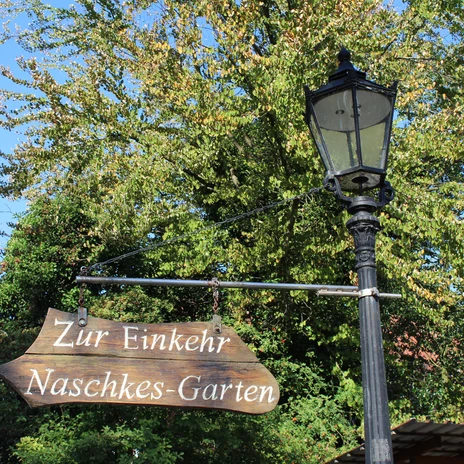 Naschkes Garten.jpg Holzschild mit der Aufschrift „Zur Einkehr Naschkes-Garten“ an einer alten Laterne vor Bäumen.