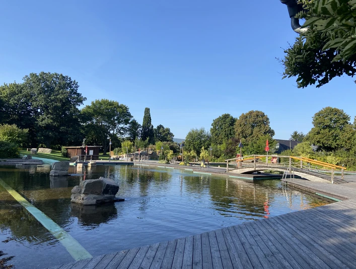 IMG_5788 4.jpeg Ein weitläufiger Park mit Teich, Holzsteg und Brücken, umgeben von vielfältigen grünen Bäumen.An extensive park with a pond, wooden walkway and bridges, surrounded by a variety of green trees.En stor park med en dam, træbroer og gangbroer, omgivet af en række grønne træer.Een uitgestrekt park met een vijver, houten loopbruggen en bruggen, omringd door een verscheidenheid aan groene bomen.