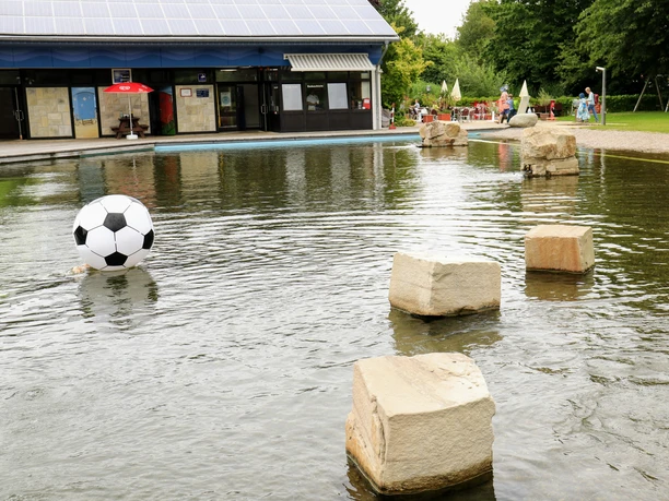 Ein Wasserbecken mit großen Steinen und einem riesigen aufblasbaren Fußball vor einem modernen Gebäude.