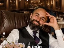 Aydın Işık sitzt lächelnd in einem eleganten Restaurant, den Kopf auf die Hand gestützt.