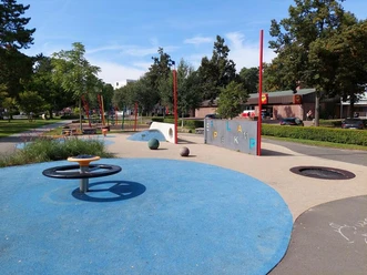 Der Spielplatz Grünanger West zeigt eine bunte, kinderfreundliche Anlage mit blauen und beigen Spielflächen. Zu sehen sind eine kreisförmige Drehscheibe, in den Boden eingelassene Trampolins und eine Kletterwand mit Buchstaben. Rote Pfosten und schattenspendende Bäume umranden den Bereich, während im Hintergrund Bänke zum Verweilen einladen. Dies ist ideal für Spiel und Erholung geeignet.