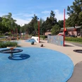 Der Spielplatz Grünanger West zeigt eine bunte, kinderfreundliche Anlage mit blauen und beigen Spielflächen. Zu sehen sind eine kreisförmige Drehscheibe, in den Boden eingelassene Trampolins und eine Kletterwand mit Buchstaben. Rote Pfosten und schattenspendende Bäume umranden den Bereich, während im Hintergrund Bänke zum Verweilen einladen. Dies ist ideal für Spiel und Erholung geeignet.