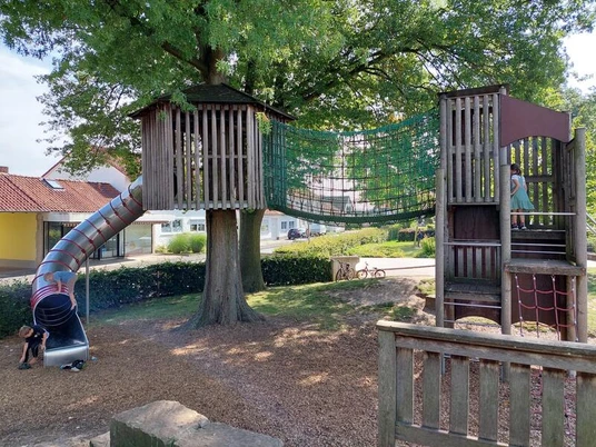 Der Spielplatz Tannenbergplatz zeigt einen einladenden Abenteuerspielplatz unter einem großen Baum. Zwei hölzerne Spielhäuser sind durch eine stabile Seilbrücke verbunden, während eine spiralförmige Rutsche von einem der Häuser hinabführt. Der Boden ist mit weichem Holzschnitzel bedeckt, was ein sicheres Spielumfeld schafft.