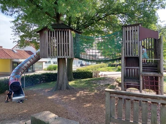 Spielplatz Tannenbergplatz Der Spielplatz Tannenbergplatz zeigt einen einladenden Abenteuerspielplatz unter einem großen Baum. Zwei hölzerne Spielhäuser sind durch eine stabile Seilbrücke verbunden, während eine spiralförmige Rutsche von einem der Häuser hinabführt. Der Boden ist mit weichem Holzschnitzel bedeckt, was ein sicheres Spielumfeld schafft.
