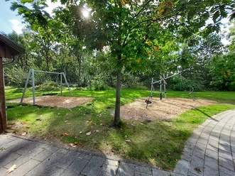 Ein Spielplatz mit zwei Schaukeln und einem Klettergerüst, umgeben von grüner Wiese und Bäumen.
