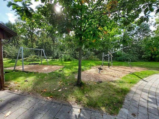 Spielplatz Jugendzentrum Isy7 Ein Spielplatz mit zwei Schaukeln und einem Klettergerüst, umgeben von grüner Wiese und Bäumen.