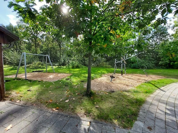 Spielplatz Jugendzentrum Isy7 Ein Spielplatz mit zwei Schaukeln und einem Klettergerüst, umgeben von grüner Wiese und Bäumen.