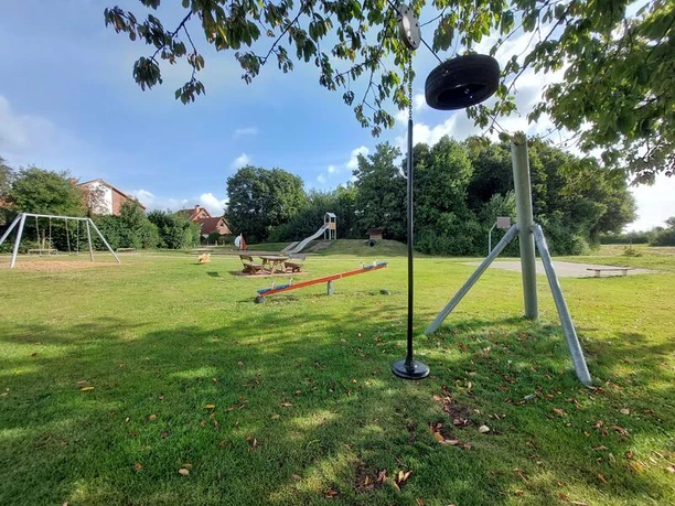 Spielplatz Frotheimer Weg Ein weitläufiger Spielplatz mit Schaukeln, Wippe und einer Rutsche auf grüner Wiese, umgeben von Bäumen.