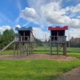 Spielplatz Rotdornstraße Der Spielplatz an der Rotdornstraße zeigt zwei hölzerne Klettertürme mit blauen und roten Dächern, verbunden durch Seile und einer Metallrutsche. Die Anlage steht auf einer Wiese unter blauem Himmel.