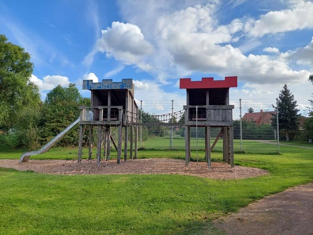 Spielplatz Rotdornstraße Der Spielplatz an der Rotdornstraße zeigt zwei hölzerne Klettertürme mit blauen und roten Dächern, verbunden durch Seile und einer Metallrutsche. Die Anlage steht auf einer Wiese unter blauem Himmel.