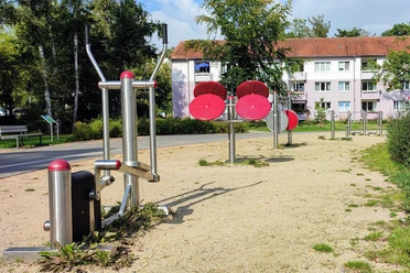 Spielplatz Boras Park Metallene Fitnessgeräte auf Kiesboden in einem Park, umrundet von Rasen und mit Bäumen im Hintergrund; im Hintergrund mehrstöckige Wohngebäude.