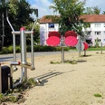 Spielplatz Boras Park Metallene Fitnessgeräte auf Kiesboden in einem Park, umrundet von Rasen und mit Bäumen im Hintergrund; im Hintergrund mehrstöckige Wohngebäude.