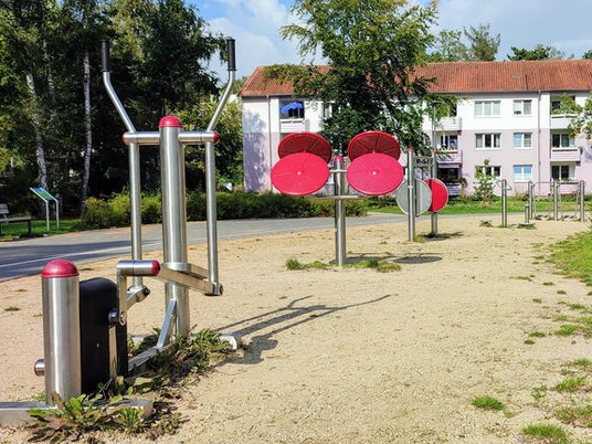 Spielplatz Boras Park Metallene Fitnessgeräte auf Kiesboden in einem Park, umrundet von Rasen und mit Bäumen im Hintergrund; im Hintergrund mehrstöckige Wohngebäude.