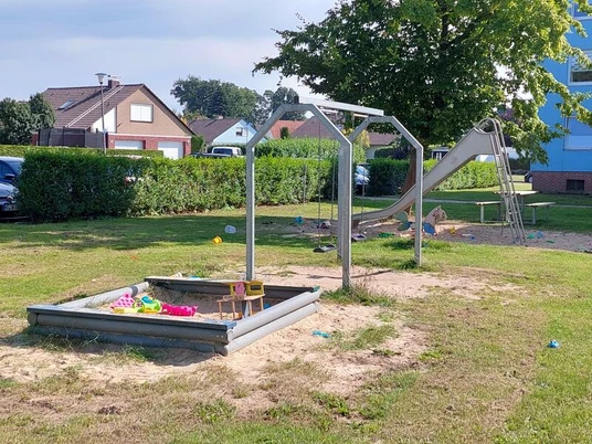 Spielplatz mit Rutsche, Sandkasten und grünem Rasen, umgeben von Wohnhäusern und Hecken.