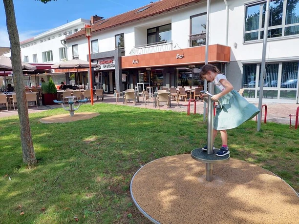 Das Bild zeigt ein Kind, das auf einem drehbaren Spielgerät auf einem Spielplatz steht, umgeben von Rasen, mit einem Gebäude im Hintergrund.