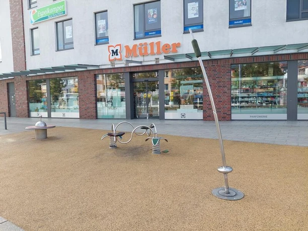 Spielplatz Breslauer Straße Der Spielplatz vor dem Gebäude mit Müller-Filiale zeigt eine moderne Schaukel und eine Wippe auf einer gepflasterten Fläche.