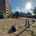 Der Spielplatz am Brandenburger Ring 16 zeigt eine Wippe aus Metall und Sand im Vordergrund, einen Kletterblock, eine Schaukel und umliegende Vegetation unter blauem Himmel.