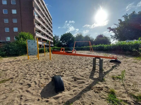 Der Spielplatz am Brandenburger Ring 16 zeigt eine Wippe aus Metall und Sand im Vordergrund, einen Kletterblock, eine Schaukel und umliegende Vegetation unter blauem Himmel.