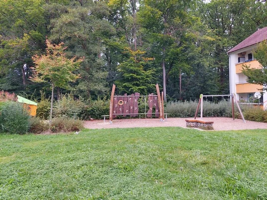Ein moderner Spielplatz in einer grünen Umgebung, umgeben von Bäumen und einem Wohngebäude. Zentral ist eine Kletteranlage mit Kletternetzen und Rutschen zu sehen, daneben eine Schaukel auf sandigem Untergrund.