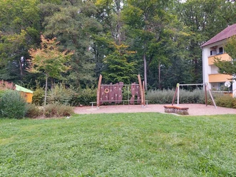 Spielplatz Brandenburger Ring 48 Ein moderner Spielplatz in einer grünen Umgebung, umgeben von Bäumen und einem Wohngebäude. Zentral ist eine Kletteranlage mit Kletternetzen und Rutschen zu sehen, daneben eine Schaukel auf sandigem Untergrund.