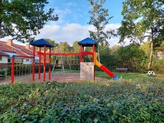 Ein farbenfroher Spielplatz mit Kletterturm, Rutsche und Kletterwand, umgeben von Bäumen und Sträuchern.
