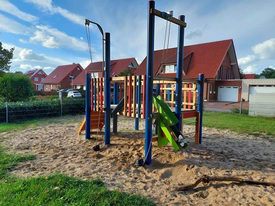 Ein farbenfrohes Klettergerüst auf einem Sandboden, umgeben von grünem Rasen und Wohnhäusern im Hintergrund.