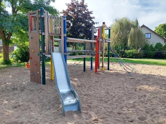 Ein moderner Spielplatz mit Klettergerüst, Rutsche und Kletterseil auf Sandboden, umgeben von Bäumen und Büschen.