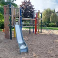 Spielplatz Espenweg Ein moderner Spielplatz mit Klettergerüst, Rutsche und Kletterseil auf Sandboden, umgeben von Bäumen und Büschen.