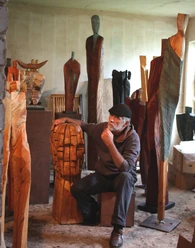 4.12_Atelierwerkstatt Rainer Ern.jpg Ein Künstler sitzt nachdenklich in seiner Werkstatt, umgeben von großen Holzskulpturen.