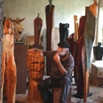 4.12_Atelierwerkstatt Rainer Ern.jpg Ein Künstler sitzt nachdenklich in seiner Werkstatt, umgeben von großen Holzskulpturen.