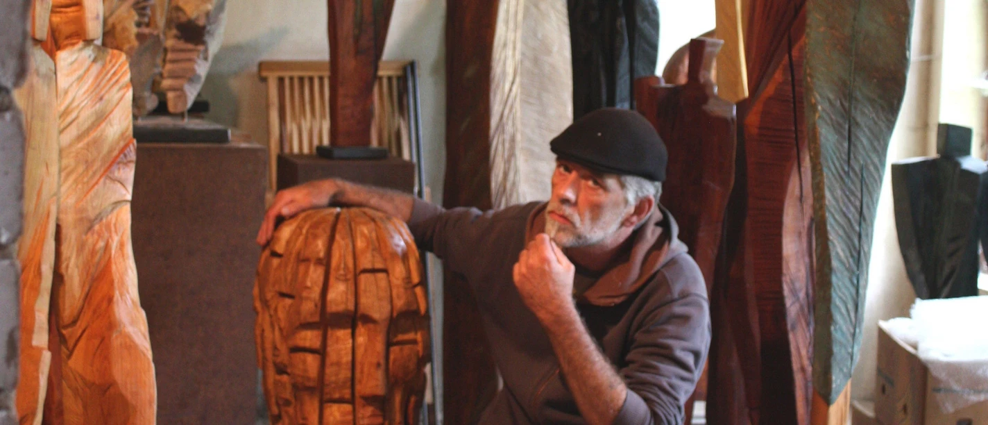 4.12_Atelierwerkstatt Rainer Ern.jpg Ein Künstler sitzt nachdenklich in seiner Werkstatt, umgeben von großen Holzskulpturen.