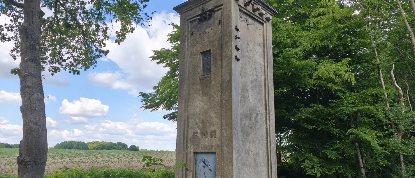 POI-1-a-Trafohaus.jpg Historisches Trafohaus in ländlicher Umgebung neben einem Wald, mit blauem Himmel und weißen Wolken.