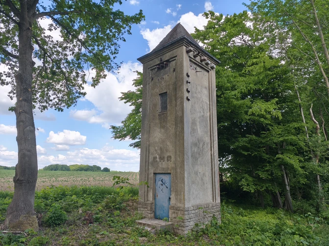 POI-1-a-Trafohaus.jpg Historisches Trafohaus in ländlicher Umgebung neben einem Wald, mit blauem Himmel und weißen Wolken.