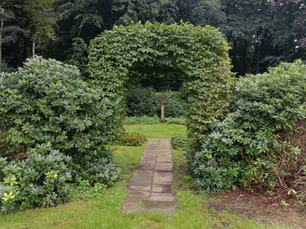 POI-3-b-Erbbegräbnis-Ebmeyer.jpg Ein Weg aus Steinplatten führt durch einen grünen, von Hecken gesäumten Durchgang zu einem hölzernen Kreuz.