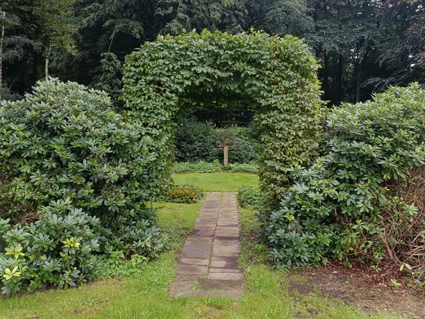 POI-3-b-Erbbegräbnis-Ebmeyer.jpg Ein Weg aus Steinplatten führt durch einen grünen, von Hecken gesäumten Durchgang zu einem hölzernen Kreuz.