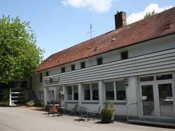 Historisches Gebäude mit Ziegelsteindach und weißen Fassadenelementen, umrahmt von Bäumen.