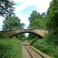 POI_0_Titelbild1-evt.JPG Steinerne Eisenbahnbrücke überspannt eine von Bäumen gesäumte Bahnstrecke in grüner Landschaft.