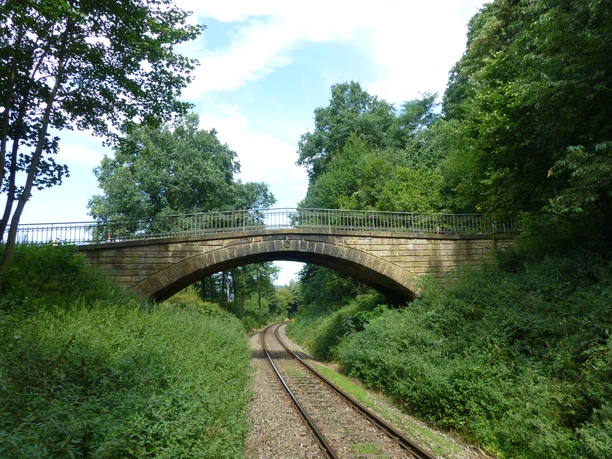 POI_0_Titelbild1-evt.JPG Steinerne Eisenbahnbrücke überspannt eine von Bäumen gesäumte Bahnstrecke in grüner Landschaft.